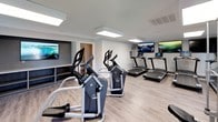The fitness center at Lake Castleton is a thoughtfully designed, single-floor facility offering a comprehensive workout environment. The space features durable wood-look flooring and neutral-toned walls accented in gray, creating a clean and modern aesthetic throughout.

The cardio area is well-stocked with multiple treadmills and elliptical machines, each equipped with individual console displays. Wall-mounted flat-screen televisions are positioned throughout the room, providing entertainment during workouts. Recessed panel lighting illuminates the space efficiently, complemented by natural light streaming through several white-trimmed windows that offer views of the surrounding greenery.

A dedicated strength training section includes a weight bench and a multi-function rack with free weights, providing options for resistance and strength workouts. Built-in shelving units along the walls offer convenient storage for personal belongings or fitness accessories.

The facility also includes a water fountain station and a separate room with mirrored walls, visible toward the rear of the space. A direct exterior exit with a windowed door adds accessibility and convenience. Overall, the fitness center delivers a functional, well-lit, and inviting environment suitable for a range of exercise routines.