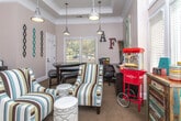 Located on Primrose Pkwy in Johns Creek, Georgia, this single-floor community space combines classic architectural charm with a warm, eclectic aesthetic. Elegant crown molding, tray ceilings, and neutral warm-toned walls establish a refined backdrop throughout, while patterned carpet flooring adds texture and continuity from room to room.

The main lounge area is a welcoming gathering spot anchored by bold multi-striped armchairs in tones of teal, brown, and cream, paired with a tufted ottoman. Industrial-style pendant lights hang overhead, complementing the casual-modern décor. A wall of tall windows floods the room with natural light and frames views of surrounding greenery, while a vintage-style popcorn machine, coffee station, and decorative accents give the space a lively, community-oriented character.

An adjoining dining nook features a dark-stained table with a leather bench, set beneath a pendant light. Decorative wall art and bold lettered signage add a playful, personalized touch to this intimate area.

A dedicated workstation room provides a functional retreat, equipped with a long desk, ergonomic chair, computer setup, and printer. A striking circular canvas artwork adorns the wall, and additional seating makes it a comfortable spot for focused work. Doorways connect the rooms, offering an open and navigable layout that balances productivity with relaxation.