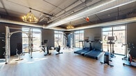 Located at 225 Canal St in Lemont, Illinois, this fitness center offers a thoughtfully designed exercise space that blends industrial aesthetics with modern functionality. The single-floor layout provides a generous, open workout area bathed in natural light through large floor-to-ceiling windows that frame views of the surrounding landscape.

The space features an industrial-chic design with exposed ductwork and piping overhead, complemented by warm Edison-style chandelier lighting that adds an inviting ambiance. Light hardwood flooring runs throughout the main workout area, creating a clean and contemporary feel.

The exercise room is well-appointed with a variety of cardio equipment, including multiple treadmills, stationary bikes, and elliptical machines. A dedicated strength training zone houses a cable machine system, a rack of dumbbells, and a bench press area. Full-length mirrors line one section of the wall, enhancing the sense of space and supporting proper form during workouts.

A separate glass-enclosed room provides a private area for focused training or stretching, complete with its own flooring and dumbbell storage. An open flex area with light wood flooring and decorative wall art offers additional room for yoga, stretching, or group exercise. The overall atmosphere is modern, bright, and energizing, making this fitness center a well-rounded facility for a comprehensive workout experience.