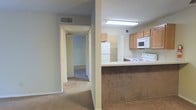This single-story space is organized around a practical, open-concept layout that connects the kitchen and living areas seamlessly. The kitchen is equipped with light oak cabinetry, a white appliance suite including a refrigerator, microwave, and electric range, and a peninsula counter finished with a brown laminate front, providing additional prep space and a natural division between the kitchen and the adjacent carpeted area. Overhead, a fluorescent light fixture ensures bright, even illumination throughout the kitchen. A hallway extends from the main living area, leading to additional rooms beyond. The hallway is carpeted in a neutral tone that continues throughout the space, contributing to a cohesive and understated aesthetic. Doors along the hallway suggest access to closets or additional rooms, and a glimpse at the far end reveals a carpeted room with natural light. The walls are finished in a soft neutral tone throughout, and a ceiling vent indicates a central HVAC system. Overall, the space presents a straightforward, functional layout suited to everyday living.