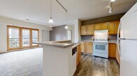 This single-floor unit is thoughtfully laid out to maximize functionality and comfort. The kitchen serves as a central highlight, featuring warm honey-toned wood cabinetry that extends from floor to ceiling, offering generous storage throughout. A peninsula with a built-in sink and dishwasher extends into the living area, creating a natural divide between the kitchen and main living space while maintaining an open, connected feel. Track lighting overhead and dual pendant lights above the peninsula provide practical and ambient illumination. White appliances, including an electric range and refrigerator, complement the neutral countertops and wood-look hard flooring found throughout the kitchen.

The spacious living area transitions seamlessly from the kitchen and is finished with plush carpeting. Warm-toned wooden French doors open to a balcony with urban views, allowing natural light to fill the room. A separate bedroom also benefits from large windows framing city views, with soft carpeting underfoot.

Two bathrooms serve the unit; one features a walk-in shower, while the other includes a full soaking tub. Both are finished with wood-toned vanities, light countertops, and wood-look flooring. Additional features include louvered closet doors and a textured ceiling throughout.