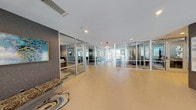 This single-floor space presents a notable contrast between two distinct areas. The main corridor is a standout architectural feature, characterized by a spacious, light-filled hallway with large-format hexagonal tile flooring in warm neutral tones. Floor-to-ceiling glass partition walls with sleek aluminum frames line both sides of the corridor, offering clear sightlines into adjacent rooms that appear to serve as meeting or office spaces. Recessed lighting runs throughout the ceiling, complementing the natural light that filters in from the far end of the hall. A decorative bench with a patterned area rug and abstract wall art add curated design touches to this professional, modern environment.

In contrast, the space also includes a compact closet area finished with neutral gray carpet and light-toned walls. This room is practical in design, equipped with white wire shelving systems mounted along the upper walls, providing organized storage capacity. A six-panel white interior door with a brushed nickel lever handle provides access. The shelving spans multiple walls, maximizing the available storage footprint. Together, these areas reflect a thoughtfully designed space that balances polished communal environments with functional utility zones.