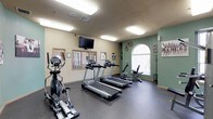 This fitness center is designed to provide a practical and comfortable environment for exercise. The space is outfitted with a variety of cardio equipment, including multiple treadmills, an elliptical machine, and a stationary bike, catering to a range of workout preferences. Durable dark flooring lines the room, providing a sturdy surface suited for physical activity.

Natural light filters in through a prominent arched window, lending an open and airy feel to the space. The walls are painted in a combination of warm beige and sage green, creating an energizing yet calming atmosphere. A wall-mounted flat-screen television is positioned for easy viewing during workouts, enhancing the overall exercise experience.

Overhead fluorescent lighting ensures the space remains well-lit throughout the day. Decorative touches, including cycling-themed artwork and athletic imagery on the walls, reinforce the fitness-focused ambiance. A coat rack near the entrance offers a convenient spot for personal belongings. Additional windows along one wall allow for further natural light, keeping the environment bright and inviting. Overall, this fitness center is thoughtfully arranged to support an active and healthy lifestyle.