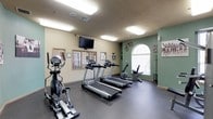 This fitness center is designed to provide a practical and comfortable environment for exercise. The space is outfitted with a variety of cardio equipment, including multiple treadmills, an elliptical machine, and a stationary bike, catering to a range of workout preferences. Durable dark flooring lines the room, providing a sturdy surface suited for physical activity.

Natural light filters in through a prominent arched window, lending an open and airy feel to the space. The walls are painted in a combination of warm beige and sage green, creating an energizing yet calming atmosphere. A wall-mounted flat-screen television is positioned for easy viewing during workouts, enhancing the overall exercise experience.

Overhead fluorescent lighting ensures the space remains well-lit throughout the day. Decorative touches, including cycling-themed artwork and athletic imagery on the walls, reinforce the fitness-focused ambiance. A coat rack near the entrance offers a convenient spot for personal belongings. Additional windows along one wall allow for further natural light, keeping the environment bright and inviting. Overall, this fitness center is thoughtfully arranged to support an active and healthy lifestyle.