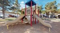 This community outdoor space offers two primary amenities designed for recreation and social gathering. At its center is a covered picnic pavilion constructed with a gabled roof supported by stone-clad columns and finished with warm wood paneling on the ceiling interior. The structure shelters a metal picnic table, providing a shaded area suitable for outdoor dining or gatherings. The open-sided design allows for natural airflow while maintaining protection from direct sunlight.

Adjacent to the pavilion is a children's playground set on a soft mulch surface, surrounded by mature trees that provide natural shade. The play structure features dual slides, a tunnel crawl-through, and interactive panels including a tic-tac-toe game, supported by red metal poles and topped with a blue canopy roof. The equipment is designed to engage children of various ages with multiple play options in a single structure.

The overall grounds are neatly maintained with concrete pathways connecting the amenity areas. Mature trees throughout the space contribute to a pleasant, park-like atmosphere. Residential buildings are visible in the background, indicating this is a shared amenity area within a larger residential community. The space is well-suited for family activities and casual outdoor enjoyment.