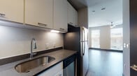 This single-floor unit showcases a clean, contemporary design with wood-look flooring and neutral tones carried throughout. The kitchen is well-appointed with stainless steel appliances, including a range, over-the-range microwave, dishwasher, and refrigerator. White cabinetry with brushed nickel hardware extends to the ceiling, providing generous storage, while dark granite-style countertops add a modern contrast. Under-cabinet lighting illuminates the workspace effectively.

The living area benefits from large windows fitted with blinds, allowing abundant natural light to fill the room. A ceiling fan with a light fixture adds both function and comfort to the space. The open layout transitions smoothly between the kitchen and living areas.

The bathroom features wood-look flooring, a white vanity with dark countertop, a framed mirror, and a tiled tub and shower combination. Overhead cabinetry above the toilet provides additional storage.

A dedicated closet area offers built-in shelving and hanging space, while a stacked washer and dryer unit is conveniently located nearby, adding in-unit laundry functionality. An additional nook adjacent to the kitchen provides flexible utility space, completing this efficiently designed unit.