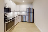 Located at 50 Barnum Ave in Port Jefferson, NY, this well-appointed single-floor unit offers a clean, modern aesthetic throughout. Warm hardwood flooring flows seamlessly across the open living and bedroom areas, complemented by crisp white walls and recessed lighting that create a bright, welcoming atmosphere.

The open-concept living area is generously proportioned, featuring crown molding and recessed lighting that add a polished finish. It connects naturally to the kitchen, which is outfitted with white shaker-style cabinetry, stone countertops, a subway tile backsplash, and a full suite of stainless steel appliances including a refrigerator, dishwasher, range, and microwave.

The bedrooms are well-sized and receive ample natural light through large windows dressed with horizontal blinds, offering views of the surrounding greenery. The clean, neutral palette of the rooms provides a versatile canvas for any furnishing style.

The bathroom is finished with classic white hex floor tile, a combined tub and shower enclosure, and brushed nickel fixtures and hardware, lending a timeless and functional design. Overall, this unit presents a cohesive, move-in-ready space with quality finishes throughout, situated in the charming community of Port Jefferson on Long Island's North Shore.