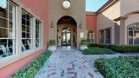 This single-story space presents a refined Mediterranean-inspired exterior, characterized by warm terracotta-toned stucco walls, a graceful arched entryway, and a decorative circular window above the entrance. Manicured boxwood hedges and potted topiaries flank the double glass doors, while a herringbone-patterned brick pathway leads visitors to the covered portico, setting an inviting and polished first impression.

Inside, the interior is defined by soaring coffered ceilings with rich dark wood detailing and an ornate chandelier that anchors the central gathering area. Natural light filters generously through a large arched transom window and additional flanking windows, illuminating the travertine-style tile flooring below. A collection of leather and upholstered armchairs surrounds glass-topped coffee tables, creating a comfortable and stylish lounge setting.

Toward the rear of the space, a vibrant teal accent wall provides a bold visual focal point, complementing a wooden reception or business desk area outfitted with cabinetry and decorative artwork. The overall ambiance balances warmth and sophistication, blending classic architectural details with contemporary furnishings for an elegant, functional environment.