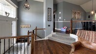 Located in Riverside, Missouri, this inviting living space showcases a thoughtfully designed interior with a warm, transitional aesthetic. Rich, dark hardwood flooring runs throughout, anchoring the room with a sense of warmth and sophistication. The space is highlighted by a stunning corner fireplace framed in stacked stone with a crisp white wood mantle, serving as a natural focal point. Decorative touches on the mantle, including vases and botanicals, add a personalized charm.

The room benefits from generous natural light streaming through tall windows fitted with classic white plantation shutters, offering both privacy and style. Cool blue-gray walls lend a calm, modern tone that complements the wood elements throughout. A geometric black pendant light fixture adds an industrial contrast to the overall design.

A carpeted staircase with elegant wood and wrought iron railings connects to additional levels of the home, flanked by decorative wall art and sconce lighting. The lofted upper landing area is open and airy, enhanced by vaulted ceilings that amplify the sense of space. This well-appointed living area balances classic architectural details with contemporary décor, creating a functional and visually appealing environment.