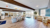 Located at 500 N Henry in historic Williamsburg, Virginia, this generously sized single-floor space offers an elegant and welcoming living room environment. The interior is defined by striking exposed dark wood ceiling beams that contrast beautifully against crisp white ceilings, lending a refined yet warm architectural character throughout. Wide-plank hardwood-style flooring runs the length of the space, adding warmth and continuity underfoot. The walls feature a classic wainscoting treatment in a soft blue-gray tone, complementing the neutral, sophisticated color palette. Natural light floods the interior through multiple large French doors and windows, which also provide direct views of and access to an outdoor patio area. The layout is open and expansive, accommodating multiple seating arrangements including woven lounge chairs, plush sofas, and accent tables that create distinct conversation areas. Elegant flush-mount and chandelier-style light fixtures, paired with wall-mounted sconces, ensure well-distributed ambient lighting. A credenza with decorative lamps and artwork anchors one wall, while potted indoor trees add organic freshness. Glass-paneled interior doors connect to adjacent rooms, enhancing the sense of openness. This space strikes a balance between timeless design and contemporary comfort.