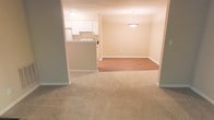This single-floor space features an open-concept layout that connects the living, dining, and kitchen areas seamlessly. The living area is finished with neutral-toned carpet, creating a comfortable and cohesive feel underfoot. Moving further into the space, the dining area transitions to hardwood-style flooring, clearly delineating the two zones while maintaining a unified aesthetic. A pendant light fixture hangs above the dining area, providing focused overhead illumination and adding a subtle decorative element to the space.

The kitchen is positioned adjacent to the dining area and is separated by a raised breakfast bar, which serves as both a functional workspace and a visual boundary. White cabinetry lines the kitchen, contributing to a clean and bright atmosphere. The walls throughout the space are painted in a warm, neutral beige tone, complementing the flooring choices and fostering an inviting ambiance. Recessed and flush-mount ceiling lighting provides consistent illumination throughout. White baseboards trim the perimeter of each area, adding a polished, finished look to the overall interior.