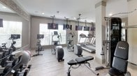 The Chestnutt's fitness center is a thoughtfully designed workout facility that balances modern aesthetics with comprehensive exercise offerings. The space features light hardwood-style flooring, crisp white walls, and structural columns that give the room a clean, contemporary feel. Exposed brick accent walls provide a warm, textural contrast to the otherwise neutral palette, while recessed lighting and ceiling fans maintain a comfortable environment throughout.

The cardio zone is well-stocked with multiple treadmills, elliptical trainers, and a stationary cycle, all positioned along large windows fitted with slatted blinds that invite natural light. Ceiling-mounted flat-screen televisions are strategically placed throughout to keep users engaged during workouts.

A dedicated strength training area includes a multi-station cable machine, an adjustable weight bench, and a full rack of dumbbells in varying sizes. Stability balls are also available for functional training. Full-length mirrors expand the sense of space and allow users to monitor their form.

A separate studio or flex space is enclosed by a frosted glass sliding door, offering a private area for stretching, yoga, or focused training sessions. Together, these zones create a versatile, well-rounded fitness facility suited to a wide range of workout routines.