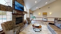 This single-story community clubhouse offers a thoughtfully designed layout encompassing multiple functional areas united by warm wood-look flooring and rich mahogany trim. The centerpiece of the main lounge is a striking floor-to-ceiling stacked stone fireplace topped with a flat-screen TV, flanked by arched windows that flood the space with natural light. Soaring vaulted ceilings amplify the open, welcoming atmosphere, while comfortable seating arrangements encourage relaxation.

Adjacent to the lounge, a casual dining area features bar-height counter seating along a granite-topped ledge and a standard dining table with chairs, with direct access to the outdoors through glass-paned doors. Wall sconces and curated artwork add a polished finishing touch throughout.

The galley-style kitchen is outfitted with rich dark wood cabinetry, granite countertops, stainless steel appliances including a refrigerator and microwave, and a double sink — well-suited for community gatherings and events.

The fitness facilities are divided into two distinct rooms: one dedicated to cardio, featuring treadmills, ellipticals, and wall-mounted TVs amid teal accent walls and mirrored surfaces; and a separate strength training room with bold orange walls, a cable machine, adjustable bench, and motivational wall decor.