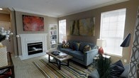 This single-floor space offers a thoughtfully designed living area that balances comfort and style. The living room serves as the centerpiece, anchored by a classic white-framed fireplace that adds warmth and a focal point to the room. Built-in shelving flanks the fireplace, providing both display and storage space, while crown molding along the ceiling adds an elegant architectural detail. Natural light pours in through multiple windows fitted with blinds, brightening the neutral taupe and warm beige walls. The living area is furnished with a plush gray sofa accented by colorful throw pillows, positioned around a wood-and-metal coffee table atop a bold, multi-colored striped area rug. Contemporary abstract artwork adorns the walls, contributing to the space's modern aesthetic. A stylish floor lamp and decorative greenery complete the room's inviting ambiance. The overall design palette blends neutral tones with vibrant pops of color through textiles and artwork, creating a dynamic yet cohesive living environment that feels both functional and visually engaging.