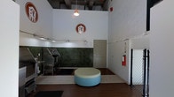 This single-floor pet care facility combines industrial and modern design elements to create a functional yet welcoming environment. The space features notably high ceilings with an exposed industrial framework, giving the interior an open, airy feel. A branded logo displayed prominently on the white-painted wall establishes the facility's identity.

The grooming area is equipped with professional stainless steel grooming stations and utility sinks, positioned against a distinctive dark mosaic tile backsplash that adds both visual interest and practical durability. Track lighting runs along the lower wall section, ensuring thorough illumination of the work area, while pendant lighting hangs from above.

The flooring transitions between tile in the grooming zone and hardwood in the waiting area, clearly delineating functional spaces. A circular, two-toned ottoman in the center of the waiting area provides seating for clients. The right side of the space features a textured white brick wall, adding architectural character. Safety equipment, including a fire extinguisher, is visibly mounted, and a secure gate is positioned near the entrance, likely for pet containment. A private door at the rear suggests additional back-of-house facilities.