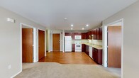 This single-floor space offers an open-concept layout that integrates the living area and kitchen into one cohesive, functional environment. The kitchen is a standout feature, outfitted with rich, dark cherry wood cabinetry that extends to the upper and lower sections of the walls, providing generous storage. White appliances — including a refrigerator, electric range, built-in microwave, and dishwasher — complement the light-toned countertops, creating a clean contrast against the warm cabinetry. A sink with under-cabinet lighting adds both practicality and ambiance to the workspace.

The flooring transitions from warm hardwood in the kitchen and entry zone to plush carpet in the living area, clearly delineating functional zones while maintaining a cohesive aesthetic. Recessed lighting throughout the ceiling provides consistent, even illumination across the open space. Multiple doors are visible, suggesting access to additional rooms or closets, as well as a main entry door with a security peephole. The overall ambiance is practical and well-organized, with a neutral color palette that lends versatility to the space.