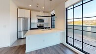 Located at 270 Waverly Ave in Mamaroneck, NY, this contemporary single-floor residence showcases a clean, modern aesthetic throughout. The open living area is defined by rich hardwood-style flooring and a striking wall of large, black-framed grid windows that flood the space with natural light and offer views of the surrounding landscape. The open-concept kitchen is thoughtfully designed with high-gloss white upper cabinetry, dark lower cabinets, a light quartz waterfall island with an integrated sink, stainless steel appliances, and a subtle glass tile backsplash. A floor-to-ceiling grid window beside the kitchen provides direct views to an outdoor terrace deck. The bedrooms continue the hardwood flooring and crisp white walls, with one bedroom featuring a full-panel glass door opening directly onto the terrace. A well-lit hallway connects the living spaces to private rooms and closets. The bathrooms are finished with spa-inspired aqua glass tile accents, including a walk-in glass-enclosed shower and a separate soaking tub with a chrome fixture suite, both set against clean white surrounds. The overall ambiance is bright, minimalist, and refined, with consistent modern detailing carried throughout every room.