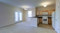 This single-floor space offers an open-concept layout that seamlessly connects the living area and kitchen. The living area features plush carpeting, neutral walls, and a chandelier centerpiece, creating a clean and inviting atmosphere. Natural light fills the space through multiple windows and a set of French doors, which also provide convenient outdoor access. The kitchen is well-equipped with light wood cabinetry, dark-toned countertops, and white appliances including a range and built-in microwave. A peninsula with matching dark countertops serves as a natural divider between the kitchen and living area, offering additional workspace and a casual dining or gathering spot. Tile flooring in the kitchen contrasts neatly with the carpeted living space, clearly defining each functional zone. The overall design is practical and functional, with a straightforward layout that maximizes usability. The neutral color palette and abundant natural light contribute to a bright, airy ambiance throughout the space.