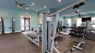 The Tracery's fitness center is a thoughtfully designed workout facility finished with soft teal-painted walls and recessed lighting that creates a bright, energizing atmosphere. The space features durable, rubberized flooring ideal for high-impact exercise and heavy equipment use. A wide selection of strength training machines, including a multi-function cable system and adjustable benches, anchors the room and provides versatile options for full-body workouts. A dedicated free weight area includes a full rack of dumbbells, catering to a range of fitness levels. Ceiling fans help maintain airflow throughout the space during intense workouts. Large mirrors along one wall visually expand the room while allowing users to monitor their form. Natural light filters in through windows and a glass-paneled entry door, offering views of the surrounding exterior. Cardio equipment is also present, rounding out the facility's offerings. The combination of natural light, modern equipment, and a well-organized layout makes this fitness center a functional and welcoming space for residents seeking an on-site exercise option at The Tracery.