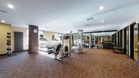 This fitness center is designed to support a full range of workout routines within a single, open floor layout. The space is finished with durable patterned carpet flooring throughout and features warm, neutral-toned walls accented by dark trim, lending the room a polished, professional feel. Recessed lighting and natural light from multiple windows combine to create a bright and energizing environment.

The cardio section is well-appointed with multiple treadmills, each equipped with individual monitor screens, alongside a stationary bike and a rowing machine — offering diverse options for aerobic exercise. The strength training area includes a multi-function cable machine with suspension training attachments, a Smith machine with a dedicated bench press setup, and a rack of free weights and dumbbells neatly organized along a mirrored wall. The mirrors extend across a large portion of the room, visually expanding the space and allowing users to monitor their form.

Additional equipment includes medicine balls, a BOSU balance trainer, exercise mats, and stability accessories, supporting flexibility and functional fitness training. A wall clock keeps track of workout time, and a water fountain station is conveniently located near the entrance. The space is well-ventilated with visible HVAC vents throughout the ceiling, ensuring a comfortable training environment.