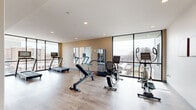 Located on the 5th floor of 239 W King St in Lancaster, PA, this modern fitness center offers a well-appointed workout space with a clean, contemporary design. The room features light hardwood-style flooring, recessed ceiling lighting, and a bright, open atmosphere enhanced by expansive floor-to-ceiling glass windows that frame sweeping panoramic views of the Lancaster cityscape and surrounding neighborhoods.

The fitness area is equipped with a variety of cardio machines, including treadmills and a stationary recumbent bike, as well as strength and conditioning equipment such as an adjustable weight bench, a rowing machine, an elliptical trainer, and free weights. A large wall-mounted mirror on one end of the room amplifies the sense of space and aids in form-checking during workouts. A colorful hula hoop and yoga block add versatility for flexibility and functional fitness routines.

A full-length glass partition wall separates the main workout floor from an adjacent area that includes restroom and changing facilities, maintaining an open, airy feel while providing privacy. Vibrant artwork adorns the white walls, adding a welcoming touch to the space. The combination of natural light, city views, modern equipment, and thoughtful layout makes this fitness center a functional and inviting amenity.