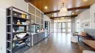 This single-floor community clubhouse is thoughtfully designed to blend modern comfort with inviting warmth. The interior is anchored by soaring wood-planked ceilings with exposed dark beams, creating a striking architectural statement throughout. Large pendant light fixtures and recessed lighting ensure the space is well-lit, complementing the abundant natural light that pours in through expansive floor-to-ceiling windows and glass-paneled French doors overlooking an outdoor pool area.

The main lounge area features a generous seating arrangement with a leather sofa, layered rugs, and a globe chandelier, offering a relaxed yet refined atmosphere. Adjacent to this is a café-style zone with built-in banquette seating, wood-topped tables, decorative throw pillows, and a mounted flat-screen TV framed by curated wall art.

A coffee and refreshment station runs along one wall, complete with gray cabinetry, open shelving stocked with décor and accessories, and a coffee machine — ideal for everyday use. The separate kitchen area is fully appointed with stainless steel appliances including a full-size refrigerator, a textured tile backsplash, dark cabinetry, open shelves, and a large communal dining table with upholstered chairs.

A media and gathering area rounds out the layout, featuring multiple wall-mounted screens, a round wood table, and additional lounge seating. Throughout, large-format tile and wood-look flooring unify the space with a clean, contemporary finish.
