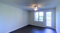 This single-story space features a clean, straightforward layout with neutral-toned walls that create a bright and open atmosphere. The main living area is finished with dark hardwood-style flooring, which provides a strong visual contrast against the light-colored walls and white baseboards, lending a polished and cohesive aesthetic throughout.

A ceiling fan with an integrated light fixture is centrally positioned overhead, offering both practical airflow and ambient lighting. Natural light flows generously into the room through a large window fitted with horizontal blinds, framing an outdoor view of lush greenery. Adjacent to the window, a glass-paneled door provides direct access to an exterior space, further enhancing the connection between the indoors and the surrounding outdoor environment.

The walls are smooth and unadorned, offering a versatile canvas for a variety of interior styles. The overall design is simple and functional, with clean lines and minimal detailing that contribute to a contemporary, move-in-ready feel. The combination of natural light, outdoor access, and durable flooring makes this a practical and comfortable living space.