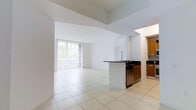 This single-floor space is defined by its airy, open-plan layout and impressively high ceilings that amplify the sense of volume throughout. The interior is finished with large-format white ceramic floor tiles, which run continuously across the living and kitchen areas, creating a cohesive and clean aesthetic.

The kitchen is positioned to one side and features warm medium-toned wood cabinetry paired with dark countertops, including a peninsula that partially separates the kitchen from the main living area. Stainless steel appliances are integrated beneath the counter, contributing a modern touch. Upper cabinetry provides practical storage while maintaining a streamlined appearance.

The living area is generously proportioned and benefits from abundant natural light streaming through a wide floor-to-ceiling sliding glass door fitted with vertical blinds. Through the glass, tropical foliage and an outdoor balcony or terrace are visible, suggesting a warm, sun-drenched setting. The all-white walls and bright natural light give the entire space a fresh, open, and welcoming ambiance throughout.