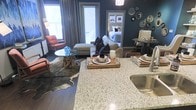 This single-floor residence showcases a thoughtfully designed open-concept layout that integrates the kitchen, dining, and living areas into one cohesive space. The kitchen features granite countertops with flecked detailing and a stainless steel sink, providing both functionality and visual appeal. A breakfast bar-style counter creates a natural transition into the living and dining zones, ideal for entertaining.

The living area is anchored by a neutral sectional sofa accented with dark throw pillows, complemented by a distinctive glass-top coffee table with a natural wood base. A cowhide-style area rug adds texture underfoot, while a retro-inspired orange accent chair introduces a bold pop of color. The deep teal walls create a dramatic, moody backdrop throughout the space, lending a curated, design-forward aesthetic.

A decorative wall arrangement of circular mirrors adds artistic character to one corner, while a small dining nook with upholstered seating sits nearby. Natural light filters in through multiple windows dressed with floor-length drapes. A partially visible bedroom through a connecting hallway reveals a neatly styled sleeping area, suggesting a well-organized floor plan throughout.