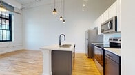 Located at 612 W Main Ave in downtown Gastonia, NC, this single-floor loft unit blends industrial character with modern finishes throughout. Warm hardwood flooring extends across the entire space, complementing the clean white walls and creating a cohesive, contemporary aesthetic.

The kitchen is a standout feature, offering a central island with a white quartz countertop, an integrated sink with a matte black faucet, and gray lower cabinetry. Full-height white upper cabinets and a complete suite of stainless steel appliances — including a refrigerator, electric range, and built-in microwave — provide both style and functionality. Soaring ceilings are accentuated by modern pendant lighting and track fixtures overhead.

The living and bedroom areas benefit from impressively tall ceilings with exposed wood beam detailing, adding to the loft's industrial charm. Generous arched and multi-pane windows framed in black allow abundant natural light to pour in while offering views of the surrounding downtown Gastonia streetscape. White-painted exposed brick walls lend authentic character to these spaces, and a ceiling fan ensures year-round comfort.

The bathroom features white shaker-style cabinetry, a white quartz vanity top, a large mirror with bar lighting, and matte black hardware accents. A laundry area is also present within the unit, adding everyday convenience to this well-appointed urban loft.