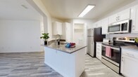 This single-floor residence is designed with a clean, modern aesthetic throughout, featuring light wood-look flooring and a crisp white color palette that creates a bright, cohesive atmosphere. The open-concept layout connects the kitchen and living area seamlessly, maximizing the sense of space and ease of movement.

The kitchen is a functional centerpiece, anchored by a peninsula island with a dark stone countertop that provides both prep space and casual seating. White shaker-style cabinetry lines the walls, offering ample storage, while stainless steel appliances — including a refrigerator, electric range, and over-the-range microwave — complete the setup. A flush-mounted ceiling light ensures the kitchen is well-illuminated. A dedicated laundry area houses a full-sized washer and dryer with overhead shelving for added convenience.

The bedroom benefits from multiple windows fitted with horizontal blinds, allowing natural light to filter through while maintaining privacy. A generously sized walk-in closet with dual hanging rods and shelving provides substantial storage capacity. The bathroom features a white vanity with stone countertop, a large mirror, and a shower-tub combination, maintaining the clean, neutral design carried throughout the space.