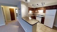 This single-floor space is designed for practical, comfortable living, with a thoughtfully laid-out kitchen serving as its focal point. The kitchen features rich, dark-stained wood cabinetry that extends to the upper walls, providing generous storage capacity. A decorative tile backsplash adds texture and visual interest behind the range and sink areas, complementing the warm tones of the cabinetry. The kitchen is equipped with a white gas range and a double-basin sink, set beneath under-cabinet lighting that brightens the workspace. Track lighting mounted to the ceiling illuminates the kitchen efficiently, while a peninsula counter with a speckled composite surface extends outward, creating a natural divide between the kitchen and the adjacent carpeted area. The tile flooring in the kitchen transitions to carpet as you move toward the hallway and connecting rooms, offering a clear delineation of functional zones. The hallway visible from the kitchen hints at additional rooms beyond, with neutral wall tones and subdued lighting maintaining a consistent, understated aesthetic throughout. Overall, the space presents a clean, functional design with classic finishes.