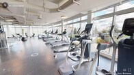 This expansive fitness facility in Durham, NC is thoughtfully designed to support a comprehensive workout experience within a bright, open single-floor layout. Floor-to-ceiling windows line the perimeter walls, flooding the space with abundant natural light and offering views of the surrounding outdoors, creating an energizing atmosphere for exercise. The high ceilings with exposed ductwork and pendant lighting contribute to an airy, industrial-modern aesthetic throughout.

The gym is generously equipped with a wide array of cardio machines, including rows of treadmills, ellipticals, stationary bikes, rowing machines, and spinning cycles, all arranged to take full advantage of the natural light and outdoor views. A dedicated strength training zone features cable machines, free weights, weight racks, benches, and multi-functional resistance equipment, providing options for both beginners and advanced users.

A standout feature is the bold, colorful geometric mural adorning one of the interior walls, adding a vibrant artistic focal point that energizes the space. Durable rubber flooring throughout ensures safety and comfort during workouts. Mirrors along key wall sections enhance the sense of openness and allow users to monitor their form. The overall ambiance is clean, modern, and motivating, making this facility a well-equipped destination for fitness in Durham.