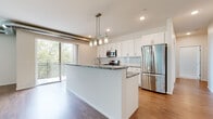 This single-floor residence showcases a contemporary design with an open-concept layout that seamlessly connects the kitchen and living areas. The kitchen serves as the centerpiece, featuring a large island topped with striking green-veined granite countertops, white shaker-style cabinetry, a light glass subway tile backsplash, and a full suite of stainless steel appliances including a gas range, built-in microwave, and refrigerator. A modern multi-pendant light fixture hangs above the island, while recessed lighting and exposed ductwork add an industrial-modern touch. Sliding glass doors lead to a private balcony with wooded views, flooding the space with natural light. The hardwood-style flooring extends throughout the main living areas, transitioning to plush carpet in the bedrooms, each of which features window blinds framing greenery views. Two bathrooms are finished in crisp white, with the primary bath offering a dual-sink vanity with ample cabinetry and a large mirror. A dedicated laundry room provides a full-size washer and dryer alongside wire shelving for storage, completing this well-appointed residence.