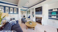 Located at 5701 Quiet Pine Circle in Chester, Virginia, this single-story community clubhouse is thoughtfully designed with a cohesive navy blue and soft aqua color palette, white wainscoting, and crown molding throughout. The grand entryway welcomes visitors with large-format tile flooring, decorative wall accents, a chandelier, and abundant natural light streaming through French doors and transom windows. The central lounge area features a coffered, beadboard ceiling, a classic white-mantled fireplace with a mounted flat-screen TV above, built-in shelving, and comfortable seating arranged around a round wood coffee table. French doors in the lounge open to views of the outdoor pool area. A dedicated business center offers built-in desks, computer workstations, and intimate seating nooks with navy half-wall accents and soft natural light from oversized windows. The fitness center is expansive and bright, with arched windows providing pool views, a coffered tile ceiling with ceiling fans, cardio equipment including treadmills, ellipticals, and a stair climber, plus a separate strength and free-weight room with floor-to-ceiling mirrors and herringbone-patterned flooring. A kitchenette area with shiplap walls, granite countertops, white cabinetry, and stainless steel appliances rounds out the amenities of this versatile community facility.