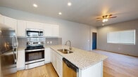 Located in Colorado Springs, CO, this single-story home offers a well-designed layout that balances functionality with modern finishes throughout. The open-concept kitchen and living area serves as the heart of the home, where light wood-style flooring flows seamlessly underfoot and a ceiling fan provides year-round comfort. The kitchen is outfitted with crisp white cabinetry, a grey tile backsplash, and granite countertops that extend across a central island featuring a undermount sink with a brushed gold faucet. Stainless steel appliances, including a refrigerator, electric range, microwave, and dishwasher, complete the space. The living area is bright and open, benefiting from natural light through a window with blinds, a wall-mounted mini-split unit for climate control, and a ceiling fan overhead. The bedroom features soft carpeting, a closet with built-in shelving behind a distinctive blue-painted paneled door, a window allowing natural light in, and a wall-mounted mini-split unit. The bathroom includes a granite-topped vanity with white cabinetry, a tub-shower combination, and wood-style flooring. A dedicated laundry nook houses a stacked washer and dryer, adding practical convenience to the home.