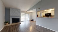 This two-bedroom, one-bathroom residence is spread across two floors, offering a well-organized layout with a dedicated entrance, closet, and combined kitchen and living area. The main living space immediately draws attention with its vaulted, angled ceiling that adds a sense of vertical openness, complemented by a textured finish overhead. A standout feature is the chevron-patterned accent wall framing a wood-burning fireplace, which serves as the focal point of the living room. Wide-plank laminate flooring runs throughout, providing a warm, cohesive aesthetic. A sliding glass door floods the space with natural light and opens to a wooden balcony overlooking mature oak trees and a quiet street below.

The kitchen is efficiently designed in a galley style, equipped with white cabinetry, granite-style countertops, a mosaic tile backsplash, and a full suite of black appliances. The bedrooms feature the same durable laminate flooring and tall ceilings, offering comfortable proportions. The primary bedroom is notably spacious and includes access to a walk-in closet with built-in wire shelving. The bathroom is clean and functional, outfitted with a tiled shower-tub combo, a single-sink vanity with a granite-style countertop, and a framed mirror.