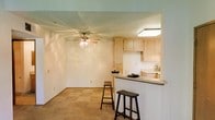 This single-story space showcases an open-concept design that seamlessly connects the living area and kitchen. The living area features neutral-toned carpet flooring and a ceiling fan with integrated lighting, lending both functionality and a casual ambiance to the room. Smooth, light-colored walls contribute to a bright and airy feel throughout the space.

A peninsula-style breakfast bar serves as the dividing element between the living area and kitchen, offering casual seating with bar stools and a practical countertop surface. The kitchen is appointed with light wood cabinetry that extends to the upper walls, providing ample storage. A flush-mounted ceiling light illuminates the kitchen workspace, complementing the natural brightness of the cabinetry. The overall color palette — consisting of warm whites, beiges, and light wood tones — creates a cohesive and inviting atmosphere. The layout is efficient and well-suited for everyday living, with the open sightlines between rooms enhancing the sense of space throughout the unit.