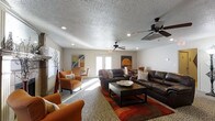 This single-floor space is designed for comfort and casual gathering, offering an open and inviting layout that encourages relaxation and social interaction. The main living area is anchored by rich, dark brown leather sofas and complemented by warm-toned upholstered armchairs in burnt orange, creating a cohesive and cozy color palette. A rectangular wood-framed coffee table serves as a central focal point, sitting atop a boldly patterned area rug that adds visual interest to the space.

The room benefits from generous natural light streaming in through a set of French doors, which also provide an access point to the exterior. Recessed lighting and dual ceiling fans are installed throughout, ensuring the space remains well-lit and comfortable year-round. The textured popcorn ceiling adds a classic character to the interior.

Decorative artwork in earthy and abstract tones adorns the walls, enhancing the warm ambiance of the space. A dining or multi-purpose table with chairs is visible in the background, offering additional functionality. The combination of patterned flooring, layered rugs, and thoughtful furnishings gives this space a practical yet stylish character suited for a variety of uses.