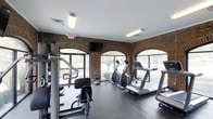 This fitness center offers a well-rounded exercise environment designed to support a variety of workout routines. The space is defined by its distinctive exposed brick walls, which lend a classic, sturdy character to the room. A series of large arched windows line the walls, flooding the interior with natural light and providing pleasant views of the surrounding greenery outdoors, creating an inviting atmosphere for exercise. The flooring consists of durable, dark rubberized material suited for high-traffic athletic use. Cardio equipment is prominently featured, including multiple treadmills and elliptical machines positioned to take advantage of the natural light from the windows. A multi-station strength training machine is also present, offering options for resistance and weight-based workouts. A wall-mounted flat-screen television is positioned for easy viewing during workouts. Overhead fluorescent lighting supplements the natural light, ensuring the space remains well-lit throughout the day. The single-floor layout keeps everything accessible and organized, making the fitness center practical and easy to navigate for users of all fitness levels.