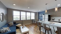 This well-appointed single-floor unit offers a thoughtfully designed open-concept layout that seamlessly connects the living, dining, and kitchen areas. Large horizontal windows flood the living space with abundant natural light while framing scenic outdoor views of surrounding greenery and neighboring rooftops. The living area features warm hardwood-style flooring, neutral gray walls accented by a bold blue feature wall in the kitchen zone, and recessed lighting throughout.

The kitchen is efficiently laid out with dark wood cabinetry, white quartz countertops, a white subway tile backsplash, stainless steel appliances including a gas range and refrigerator, and stylish pendant lighting overhead. A breakfast bar provides additional seating and casual dining space.

The bedroom is a comfortable retreat with soft neutral tones, a vibrant yellow accent wall, a closet with built-in shelving, and a window offering natural light and outdoor views. A second sleeping area features similar hardwood-style floors and window access.

The bathroom is finished with clean white subway tile extending from floor to ceiling around the tub and shower, complemented by a floating wood-toned vanity, a round mirror, and brushed nickel fixtures — creating a crisp, modern aesthetic throughout.