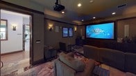 This private movie theater is designed to deliver an immersive cinematic experience within an intimate, thoughtfully appointed setting. The room is anchored by a large projection screen mounted within a dark wood built-in cabinet wall, complemented by a ceiling-mounted projector that delivers a vivid, theater-quality picture. Recessed ceiling lighting and wall-mounted sconces provide warm, dimmable ambient illumination, enhancing the cinematic atmosphere without overwhelming the space.

Seating is arranged across multiple rows, consisting of plush sofas and upholstered armchairs in earthy tones, offering comfortable viewing angles from every position. A decorative area rug with a bold floral motif covers the floor, adding visual warmth to the room. Classic framed movie posters—including recognizable titles such as Casablanca and Giant—line the side walls, reinforcing the cinematic theme with a nod to Hollywood's golden era.

Wall-mounted surround sound speakers are positioned throughout for an enveloping audio experience. Dark wood trim along the ceiling line and cabinetry provides refined architectural detail. The overall ambiance is cozy and deliberately theatrical, balancing the energy of a true movie house with the comfort of a private screening room.