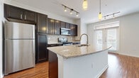 This single-floor residence offers a thoughtfully designed layout combining modern finishes with functional living spaces throughout. The kitchen serves as a central highlight, showcasing rich dark-espresso cabinetry, granite countertops, and a full suite of stainless steel appliances including a refrigerator, range, and built-in microwave. A decorative mosaic tile backsplash adds texture and visual interest, while a spacious peninsula with an integrated sink and pendant lighting above provides additional prep space and casual seating. Track lighting and hardwood flooring complete the kitchen's polished aesthetic, which flows seamlessly into an adjacent living area brightened by a large window with exterior views. The bedrooms feature neutral-toned carpet and windows that invite natural light, creating a calm and comfortable atmosphere. One bedroom includes a ceiling fan for added comfort, while another connects directly to a generously sized walk-in closet with built-in shelving and hanging rods. The bathroom is finished with large-format tile flooring, a dark-cabinetry vanity with a granite countertop and framed mirror, and a tub-shower combination with tile surround. Crown molding accents and consistent use of quality materials throughout the space lend a cohesive, refined character to the overall design.