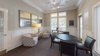 Located at 3960 Bell Rd in Nashville, Tennessee, this single-floor community space combines classic architectural design with functional, well-appointed interiors. The space is characterized by warm neutral tones, white wainscoting panels, and detailed crown molding that lend a polished, traditional aesthetic throughout.

The main lounge area serves as the heart of the space, featuring a striking coffered ceiling with elegant trim work and recessed track lighting. Generous windows with transom panes invite abundant natural light and provide views of the surrounding greenery, creating a bright and inviting atmosphere. Comfortable seating arrangements, including upholstered chairs and a round table with leather chairs, make this area ideal for both relaxed gatherings and meetings.

A dedicated workspace nook is equipped with built-in cabinetry, shelving, and a computer workstation, offering a practical area for focused tasks. An adjacent entry area with light tile flooring provides a clean, functional transition space, complete with a water dispenser for added convenience.

Throughout the space, dark patterned carpet grounds the lounge and office areas, contrasting effectively with the crisp white millwork. Multiple doorways connect the various zones seamlessly, supporting easy circulation. The overall ambiance is refined yet approachable, reflecting a thoughtfully designed environment suited for community use in the Nashville area.