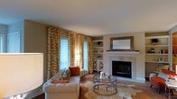 This single-floor residence offers a thoughtfully designed living space that balances comfort and contemporary style. The living room serves as the centerpiece of the home, anchored by a classic tile-surround fireplace topped with a decorative mosaic-framed mirror, creating an elegant focal point. A mounted display area above the mantle adds both function and visual interest to the space.

Built-in shelving flanks the fireplace area, providing ample display and storage space while contributing to the room's refined character. Large windows dressed in bold geometric-patterned gold and white curtains allow generous natural light to fill the room, enhancing the warm, neutral color palette of the walls and furnishings.

The living area is furnished with a tufted sofa accented with decorative pillows, complemented by a layered cowhide rug and a sleek glass-top coffee table with a metallic frame. A decorative white cabinet with lattice-patterned doors adds a stylish storage element to the room. The hardwood flooring grounds the space and ties the design elements together seamlessly.

Overall, the home exudes a warm and inviting ambiance, combining classic architectural details with modern furnishings to create a livable yet polished interior.