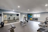 Located at 7310 Standifer Gap Rd in Chattanooga, TN, this fitness center offers a comprehensive, single-level exercise facility designed to support a wide range of workout routines. The room is finished with cool blue-gray walls, recessed lighting, and durable rubber flooring throughout, creating a clean and functional environment well-suited for physical activity.

The space is thoughtfully equipped with a variety of cardio machines, including two treadmills, an elliptical trainer, a stationary bike, and rowing machines. A dedicated free weights area features a multi-tiered dumbbell rack stocked with a full range of weights, while a storage rack along one wall holds exercise balls in multiple sizes and additional fitness accessories.

A large wall mirror runs along one side of the room, expanding the sense of space and allowing users to monitor their form during workouts. A wall-mounted flat-screen television is positioned above the mirror area, providing entertainment during cardio sessions. Adjustable weight benches offer versatility for strength training exercises.

Natural light enters through windows and a glass-paneled door, which opens to an outdoor area. Interior windows on the opposite wall provide views into an adjacent common area, further enhancing the open and connected feel of the space. The overall ambiance is bright, practical, and welcoming for daily fitness use.