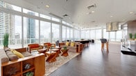 This expansive single-floor amenity space is defined by its striking floor-to-ceiling glass walls, which wrap around the perimeter and flood the interior with natural light while offering sweeping panoramic views of the surrounding urban skyline and waterway beyond. Large-format dark tile flooring grounds the open-plan layout, which is organized into several distinct functional zones that flow seamlessly into one another.

The primary lounge area features a generous seating arrangement with neutral upholstered sofas, warm-toned wooden armchairs with bold accent cushions, and sculptural live-edge wood coffee tables anchored on a geometric patterned area rug. Adjacent to this is a formal dining zone centered around a substantial solid wood communal table surrounded by dark contemporary chairs, illuminated by a cluster of pendant lights overhead.

A fully equipped kitchen with a breakfast bar, overhead ventilation hood, and brick-accented feature wall provides a social culinary hub. A dedicated game room houses a full-size billiards table alongside casual seating. The space also connects to an outdoor terrace furnished with weather-resistant lounge chairs, offering unobstructed open-air views of the cityscape and horizon.