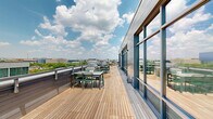Located in Washington, DC's Southwest neighborhood, this rooftop terrace offers a generously sized open-air retreat finished with warm, natural wood decking that runs the full length of the space. The deck is flanked along one side by a modern building facade clad in gray horizontal siding, punctuated by large floor-to-ceiling windows and glass doors that flood the interior with natural light while providing seamless access to the outdoors.

At the heart of the terrace's amenities is a fully equipped outdoor kitchen station, featuring a stainless steel gas grill, a built-in sink with countertop workspace, and integrated cabinetry — all designed for effortless al fresco entertaining. Adjacent to the kitchen, multiple dining tables with green chairs are thoughtfully arranged to accommodate gatherings of various sizes, while raised planters with greenery add a touch of nature to the setting.

The terrace is enclosed by a secure perimeter railing, beyond which sweeping panoramic views of the Washington, DC skyline unfold — including glimpses of landmark structures and lush tree canopies stretching into the distance. The open-sky exposure ensures abundant natural light throughout the day, making this rooftop an ideal venue for outdoor dining, social events, or simply enjoying the urban landscape.