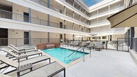 This inviting courtyard pool area in Lafayette, Louisiana, offers a well-appointed outdoor retreat nestled within the interior of a multi-story building. The space is defined by its open-air design, with the surrounding structure rising several floors and featuring balconies with metal railings that overlook the courtyard below, creating a sense of enclosure while still allowing natural light to flood the area.

At the heart of the space is a clear, well-maintained swimming pool with a charming brick water feature wall that adds a classic architectural accent. Entry points with metal handrails provide safe pool access, and the pool deck is finished in smooth, light-toned concrete that extends generously throughout the courtyard.

Surrounding the pool, a row of contemporary lounge chairs with side tables invites relaxation. A designated seating zone features multiple outdoor dining tables paired with cushioned chairs and large shade umbrellas, providing comfortable spots for gathering. The perimeter of the pool area is secured by metal fencing with a gated entry.

The building façade enclosing the courtyard is finished in neutral tones with dark accent columns, complementing the clean, modern aesthetic of the outdoor space. Overall, the area strikes a balance between functionality and comfort, making it a practical and pleasant amenity.