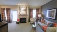 This single-floor space offers a thoughtfully designed interior that balances comfort and style. The living area serves as the centerpiece, anchored by a tile-surround fireplace topped with a large framed landscape artwork, creating a focal point that draws the eye immediately upon entry. Neutral beige and taupe tones across the walls and carpet establish a warm, inviting ambiance, while bold coral and rust accent pillows and patterned drapery add depth and visual interest. Natural light filters in through multiple windows dressed in rich, plaid curtains, as well as through a glass door that brightens the space and provides an outdoor connection. Track lighting overhead supplements the natural light, ensuring the room is well-lit at all times. The layout accommodates a comfortable seating arrangement, including a leather sofa and upholstered armchair positioned around a distinctive glass-top coffee table with a natural wood base, lending an organic, earthy touch to the otherwise refined interior. The carpeted flooring adds softness underfoot and contributes to the overall cozy atmosphere. Together, these elements create a cohesive and welcoming living environment suited for everyday comfort and relaxed entertaining.
